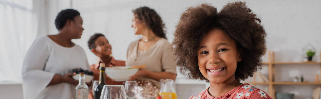 Uśmiechnięta afrykańska dziewczyna patrzy w kamerę w pobliżu rozmytej rodziny i dziękczynny obiad, baner  - Zdjęcie, obraz