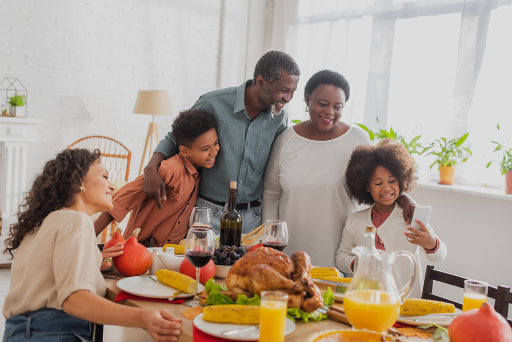 Ragazza afroamericana che tiene smartphone vicino alla famiglia e cena del Ringraziamento  - Foto, immagini