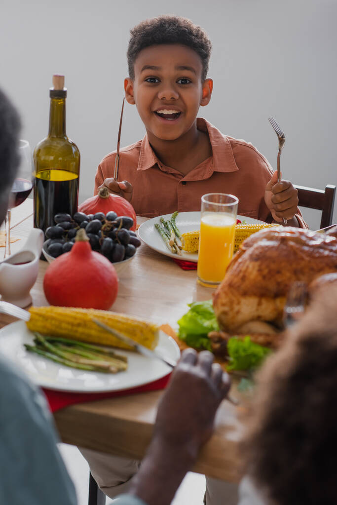 おいしい感謝祭の夕食とぼやけた家族の近くで刃物を保持している陽気なアフリカ系アメリカ人の少年  - 写真・画像