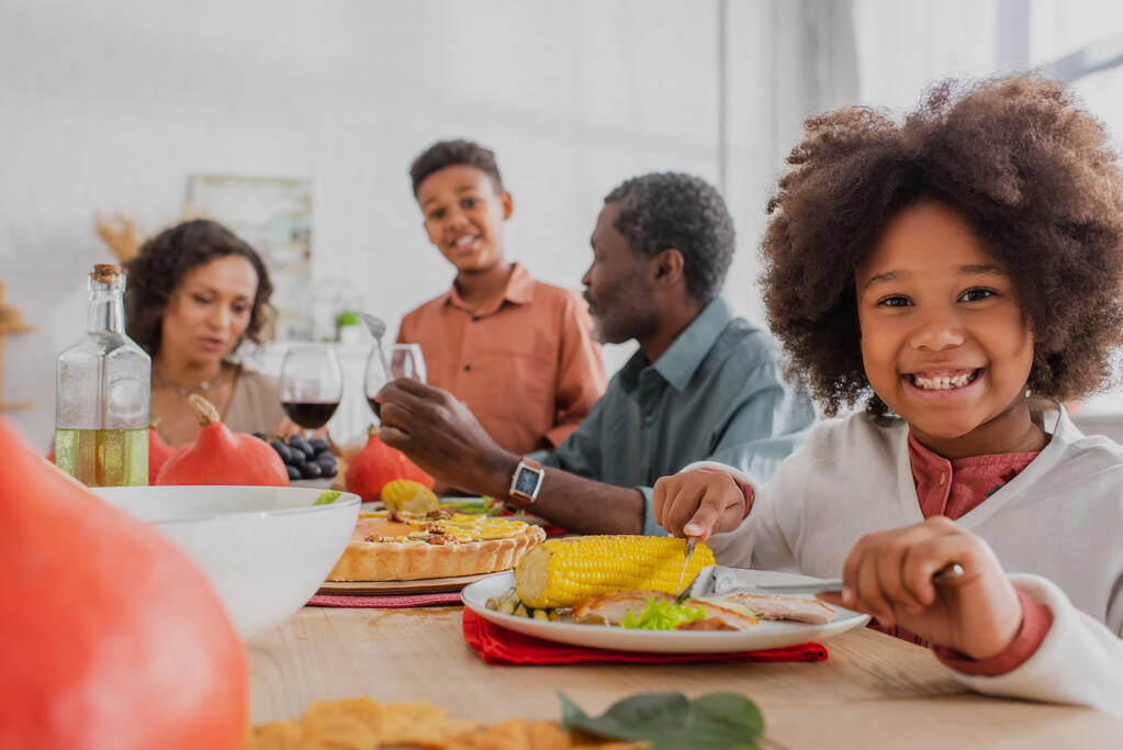 Χαρούμενη Αφροαμερικανή που κόβει καλαμπόκι κοντά σε θολή οικογένεια και δείπνο Ευχαριστιών.  - Φωτογραφία, εικόνα