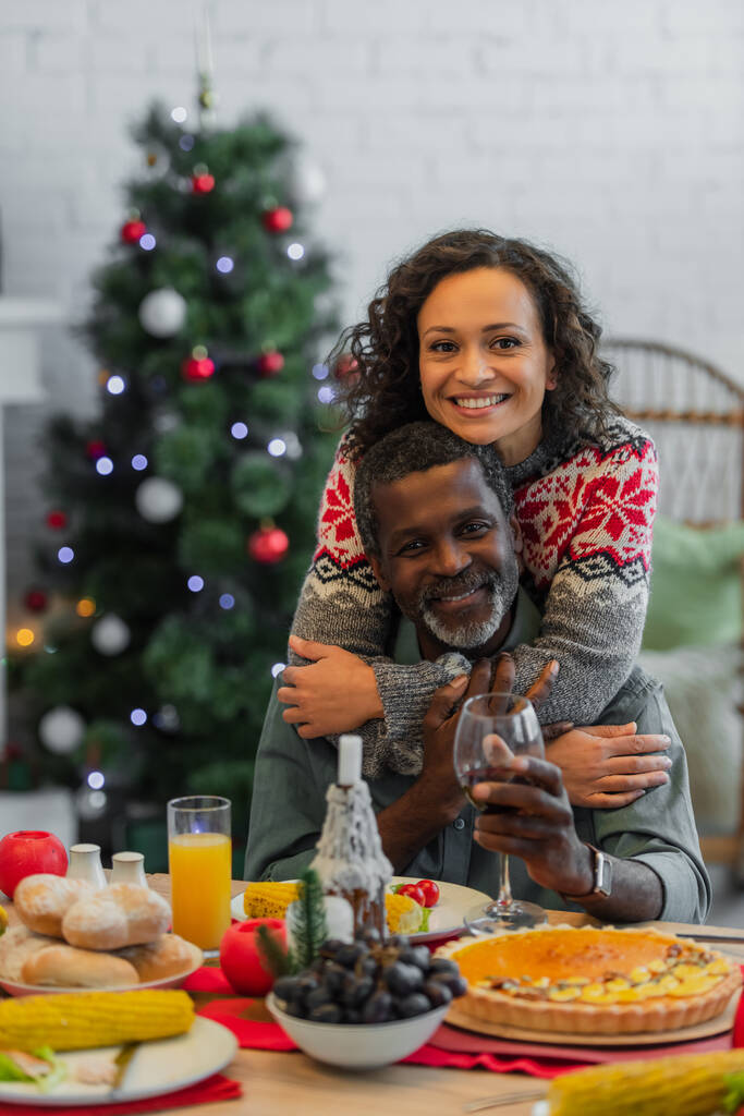 mujer afroamericana feliz abrazando papá sosteniendo copa de vino tinto durante la cena de Navidad - Foto, Imagen