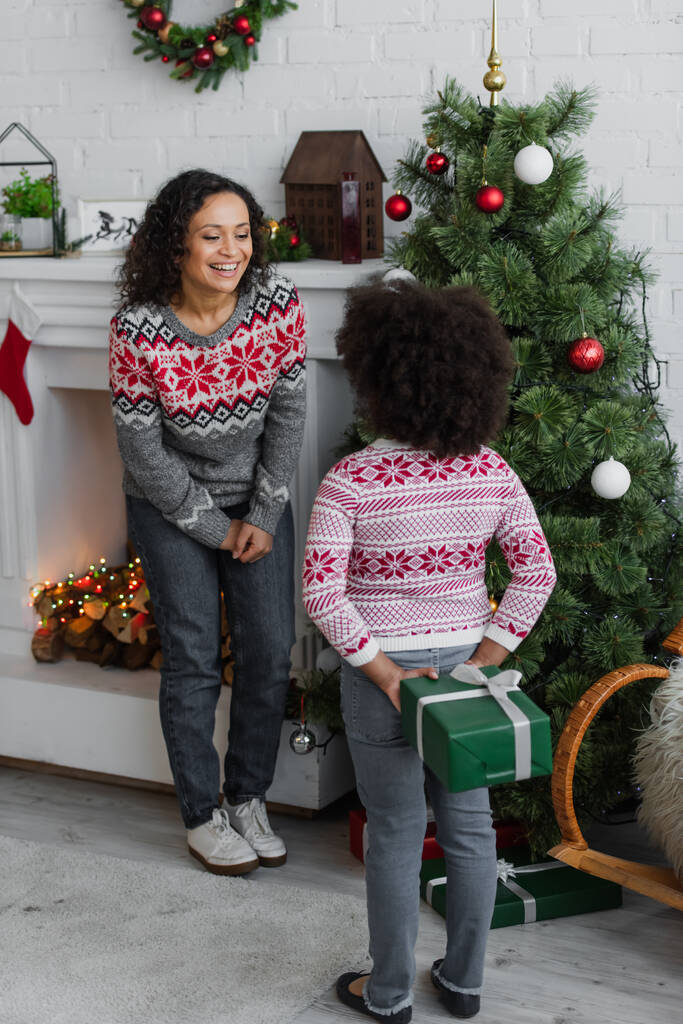 вид ззаду афроамериканська дівчина тримає подарунок на Різдво біля веселої матері
 - Фото, зображення