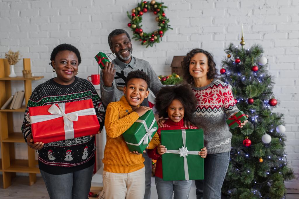 Mutlu Afro-Amerikan ailesi Noel hediyeleriyle evde kameraya bakıyor. - Fotoğraf, Görsel