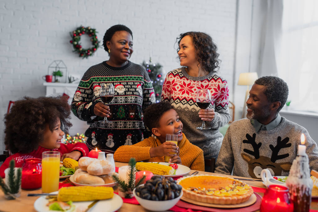 家族の近くに赤ワインのグラスを持つアフリカ系アメリカ人女性の笑顔とお祝いのディナーテーブル - 写真・画像
