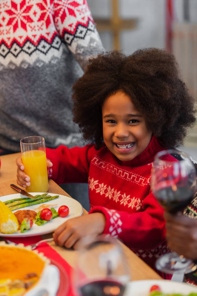 alegre afroamericana chica con jugo de naranja cerca borrosa abuelos en la cena de Navidad - Foto, imagen