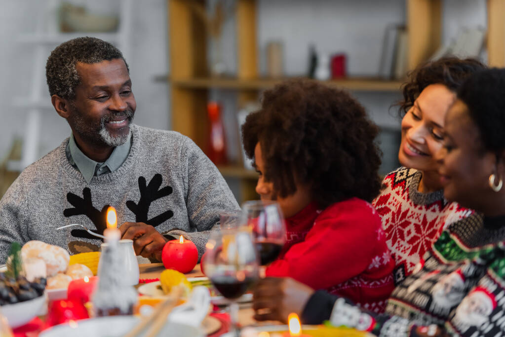 szczęśliwy Afroamerykanin człowiek patrząc na wnuczkę w pobliżu rozmytej rodziny na świąteczny obiad - Zdjęcie, obraz
