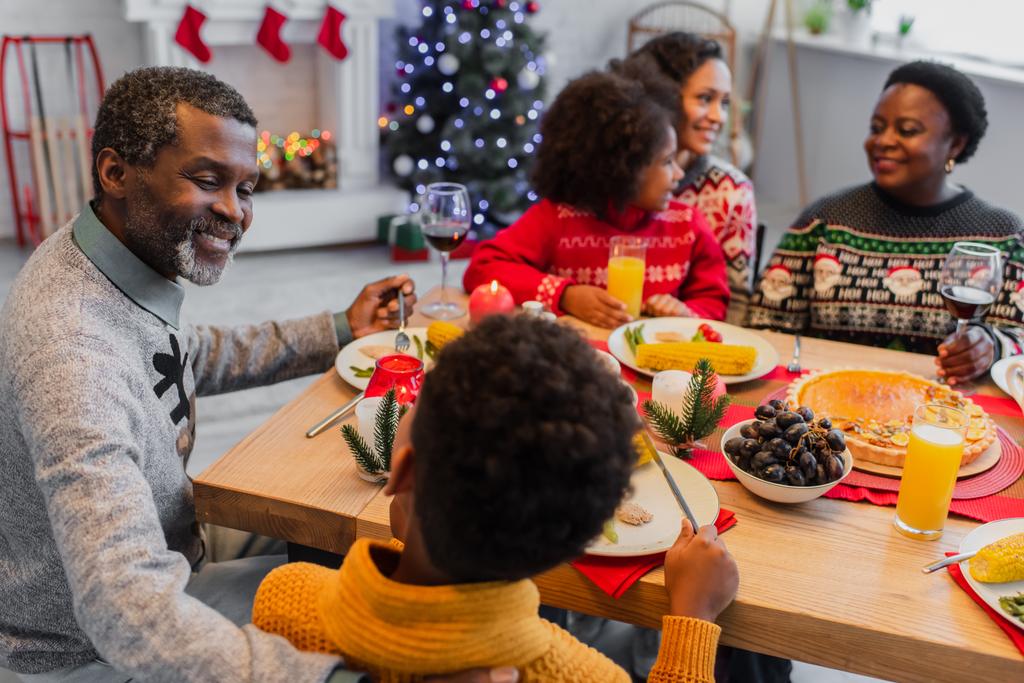 hombre afroamericano feliz hablando con el nieto cerca de la cena de Navidad con la familia borrosa - Foto, imagen