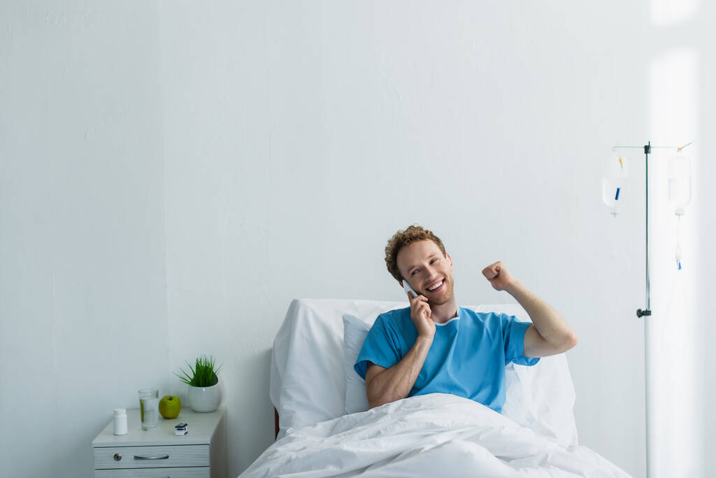 kudrnatý muž v šatech pacienta mluví na smartphonu v nemocnici  - Fotografie, Obrázek