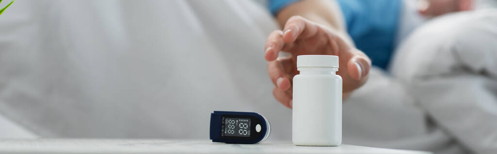 vue recadrée d'un patient flou atteignant une bouteille avec des médicaments sur une table de chevet près d'un appareil oxymètre, bannière - Photo, image