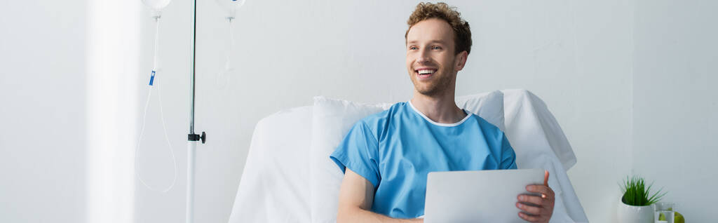 χαρούμενος ελεύθερος επαγγελματίας σε φόρεμα ασθενή χρησιμοποιώντας φορητό υπολογιστή στο κρεβάτι του νοσοκομείου, πανό - Φωτογραφία, εικόνα