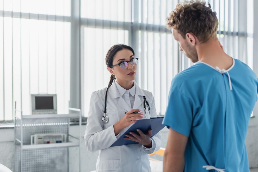 brunetka lékař s klipboard při pohledu na kudrnatý pacient v nemocnici  - Fotografie, Obrázek