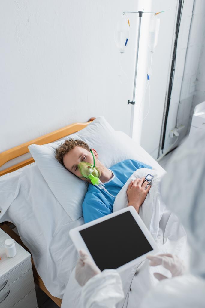 hoge hoek uitzicht van zieke patiënt in zuurstofmasker op zoek naar arts in hazmat pak en latex handschoenen met digitale tablet  - Foto, afbeelding