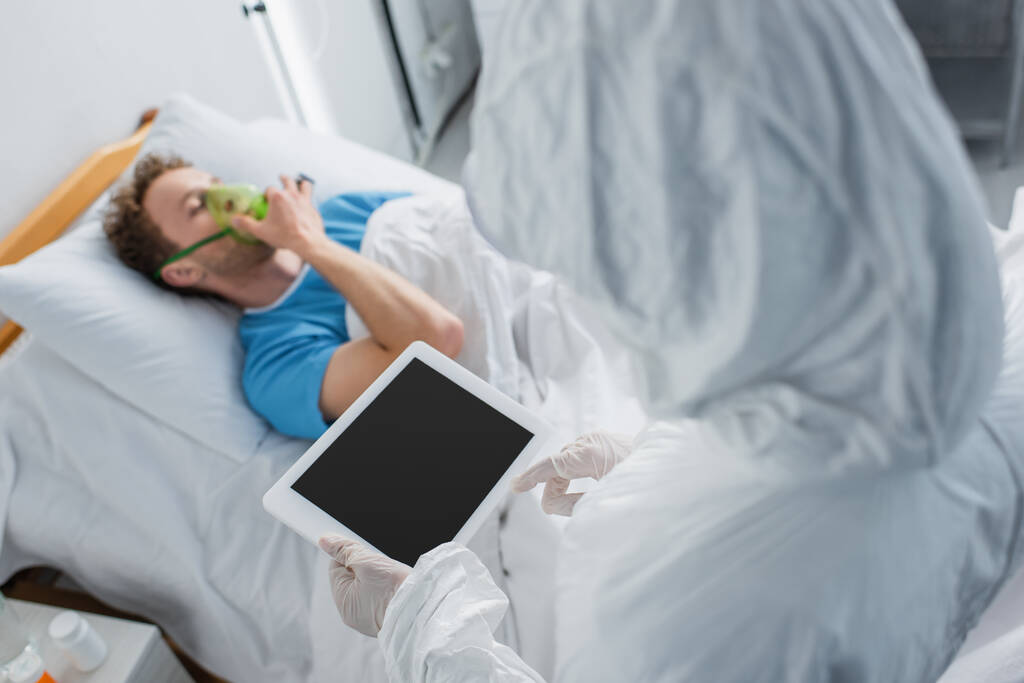 vista ad alto angolo del medico in tuta hazmat e guanti in lattice con tablet digitale con schermo bianco vicino al paziente malato  - Foto, immagini