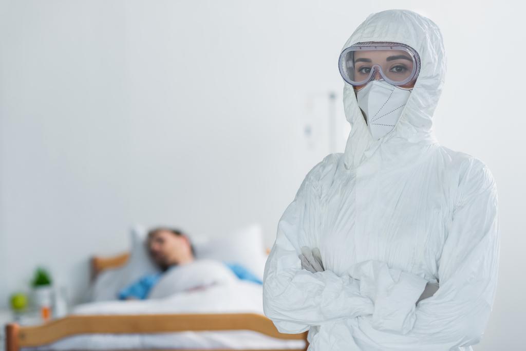 arts in persoonlijke beschermingsmiddelen en bril staan met gekruiste armen in de buurt wazig patiënt in ziekenhuis bed  - Foto, afbeelding