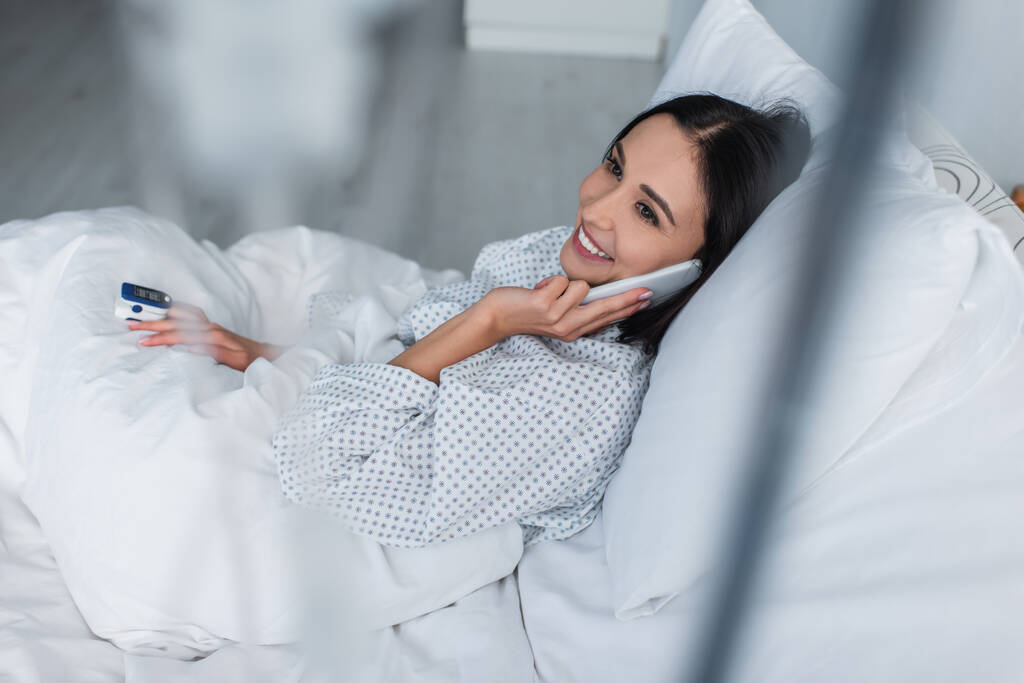 χαμογελαστή γυναίκα με οξύμετρο στο δάχτυλο που μιλάει στο smartphone ενώ βρίσκεται στο κρεβάτι του νοσοκομείου - Φωτογραφία, εικόνα
