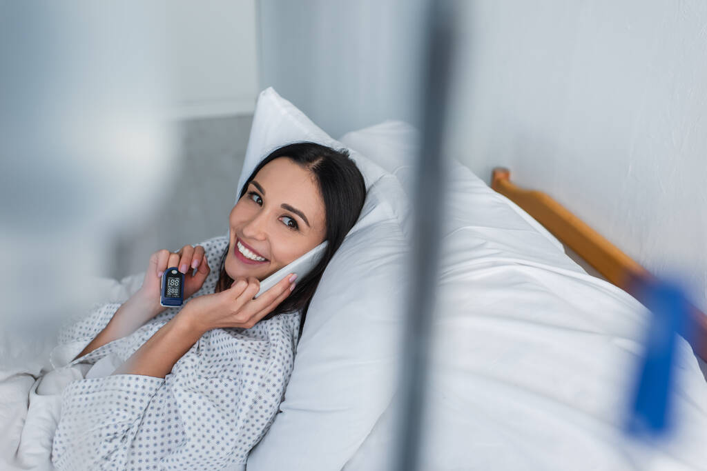 pozytywna kobieta z oksymetrem na palcu mówiąca na smartfonie leżąc na szpitalnym łóżku - Zdjęcie, obraz