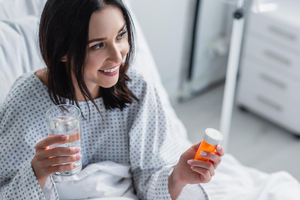 glimlachende zieke vrouw met fles met medicatie en glas water in het ziekenhuis  - Foto, afbeelding