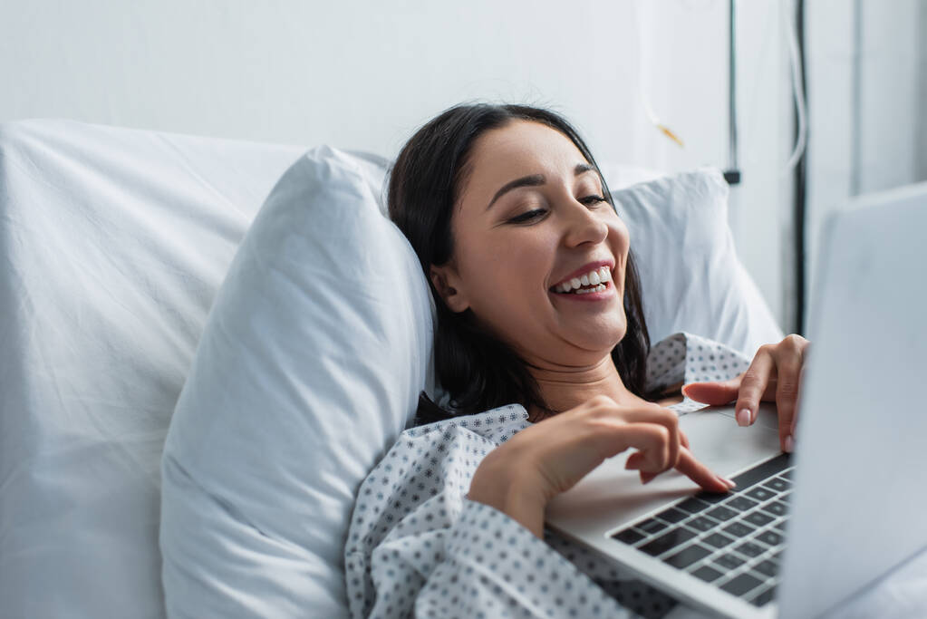 Χαρούμενη γυναίκα βλέποντας κωμική ταινία στο φορητό υπολογιστή στο κρεβάτι του νοσοκομείου - Φωτογραφία, εικόνα