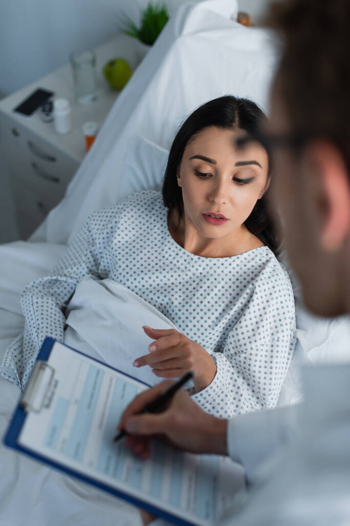 high angle view of brunette woman pointing at clipboard near blurred doctor - Photo, Image