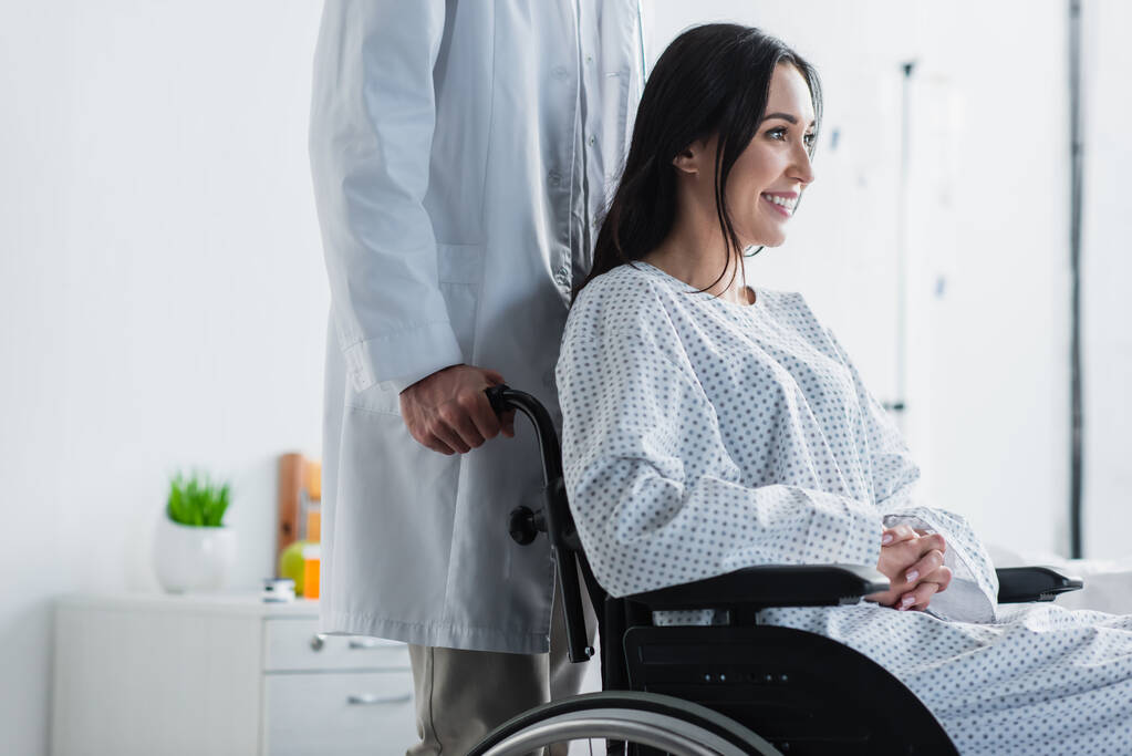 dokter in witte jas staan achter glimlachende gehandicapte vrouw in rolstoel  - Foto, afbeelding
