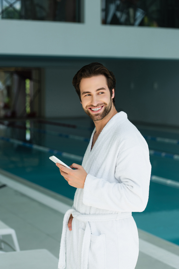 Positive man in bathrobe holding smartphone in spa center  - Photo, Image