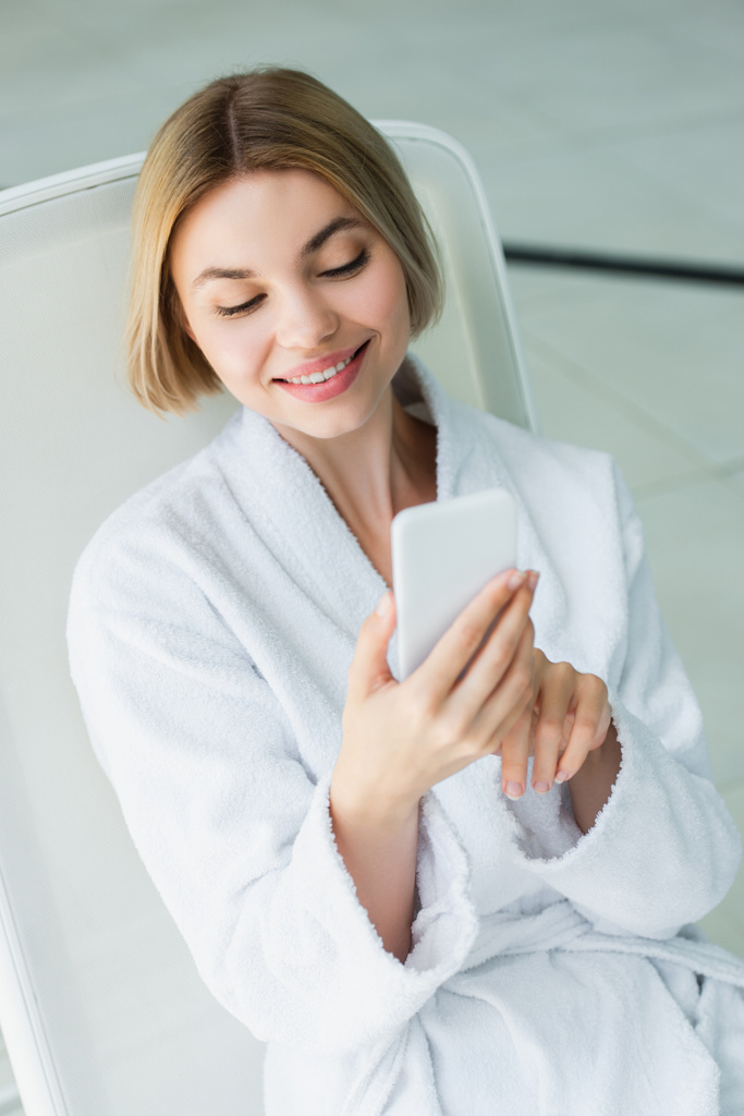 Mujer sonriente en albornoz con teléfono inteligente en la silla de cubierta en el centro de spa  - Foto, imagen