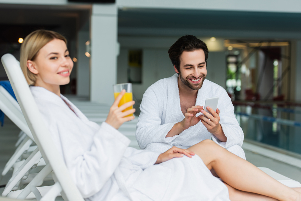 Glimlachende man in badjas met behulp van mobiele telefoon in de buurt wazig vriendin met sinaasappelsap in spa centrum  - Foto, afbeelding