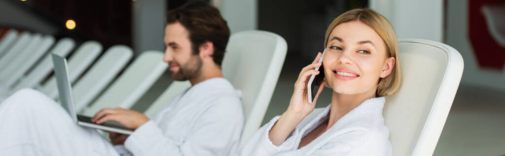 Happy woman in bathrobe talking on smartphone near blurred boyfriend with laptop in spa center, banner  - Photo, Image