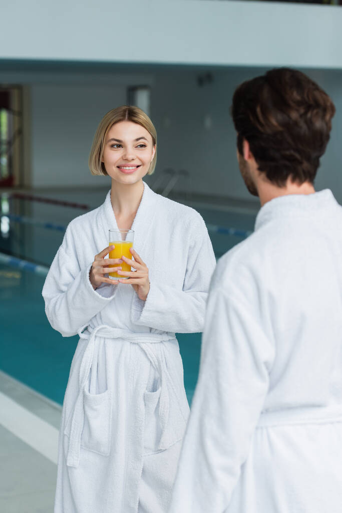 Vrolijke vrouw met een glas sinaasappelsap in de buurt van wazig vriendje in witte badjas in spa centrum  - Foto, afbeelding
