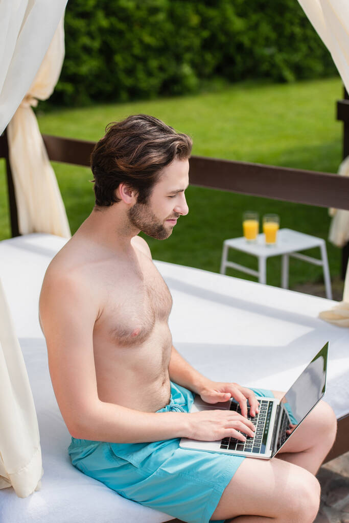 Side view of man in swimming trunks using laptop on lounge bed outdoors  - Photo, Image