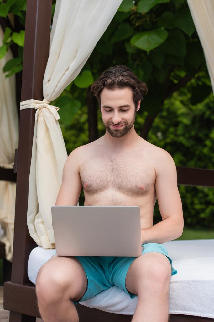 Jongeman in zwembroek met laptop op loungebad op resort  - Foto, afbeelding