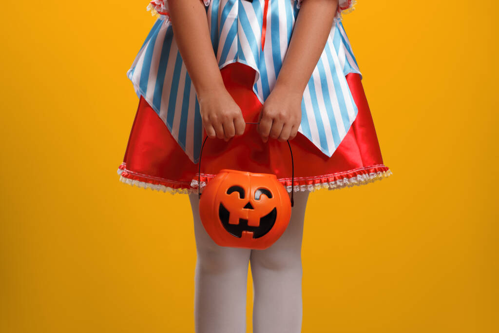Sarı arka planda Cadılar Bayramı için karnaval kostümü giymiş küçük bir kız. Çocuk elinde balkabağı feneriyle bir kova tutuyor.. - Fotoğraf, Görsel