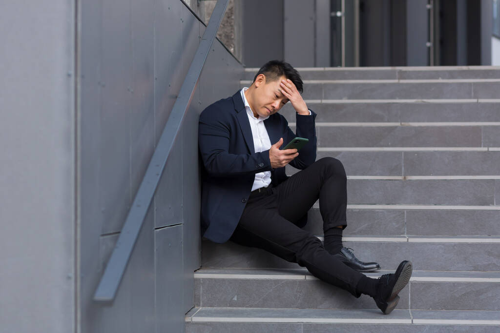 Aziatisch zakenman leest slecht nieuws van de telefoon, zitten in de buurt van het kantoor op de trap, depressief, de hoop te verliezen - Foto, afbeelding