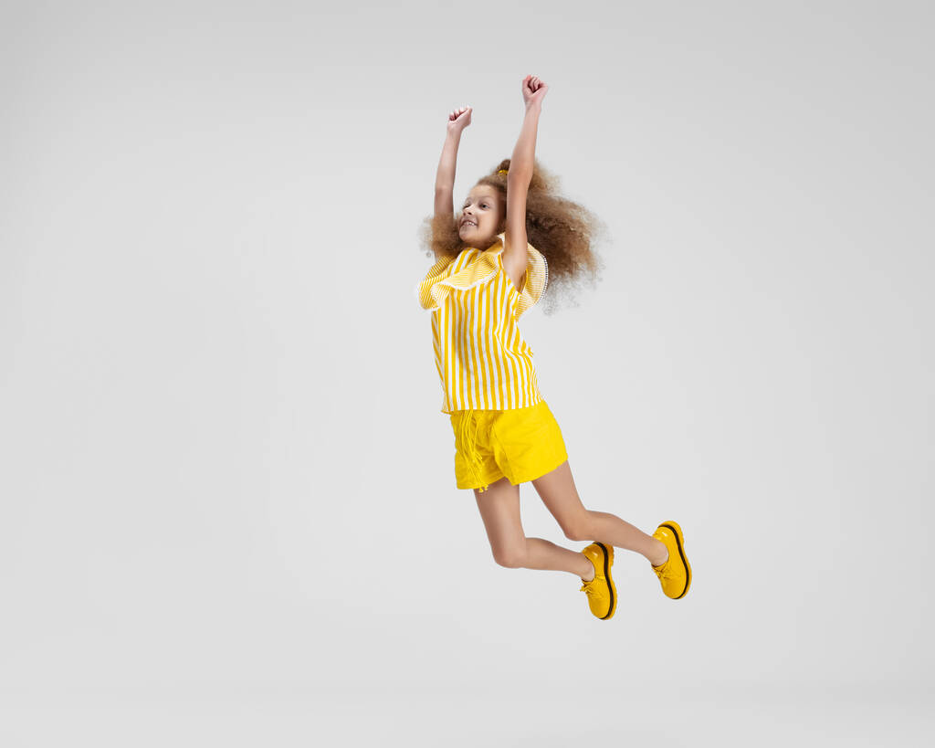 Полнометражный портрет маленькой активной кавказской дошкольницы в ярко-желтом наряде и туфлях, прыгающей на белом фоне - Фото, изображение