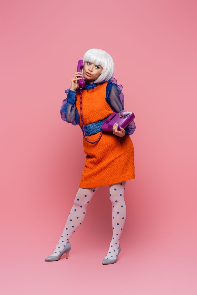 Скептическая азиатская модель поп-арта разговаривает по телефону на розовом фоне - Фото, изображение