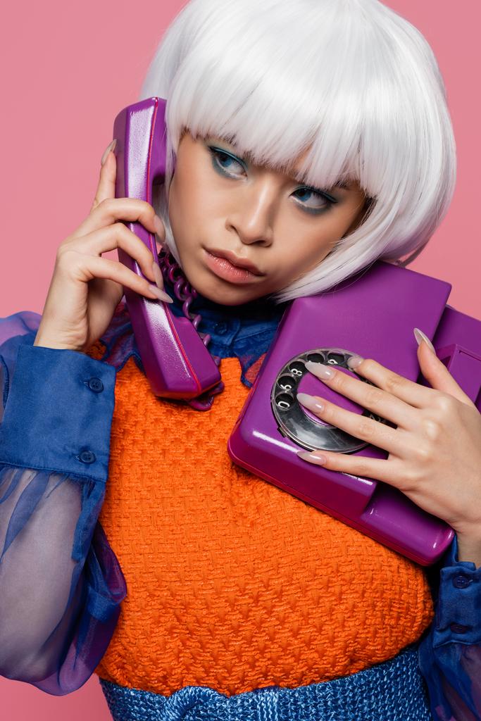 Азійська модель поп-арту в білій перуці говорить на ретро телефоні ізольованому на рожевому  - Фото, зображення