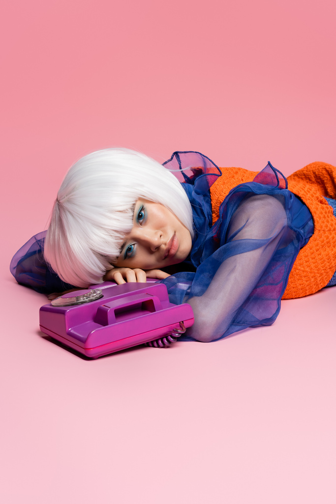 Азійська модель поп-мистецтва в білій перуці дивиться на камеру, коли лежить біля телефону на рожевому фоні  - Фото, зображення