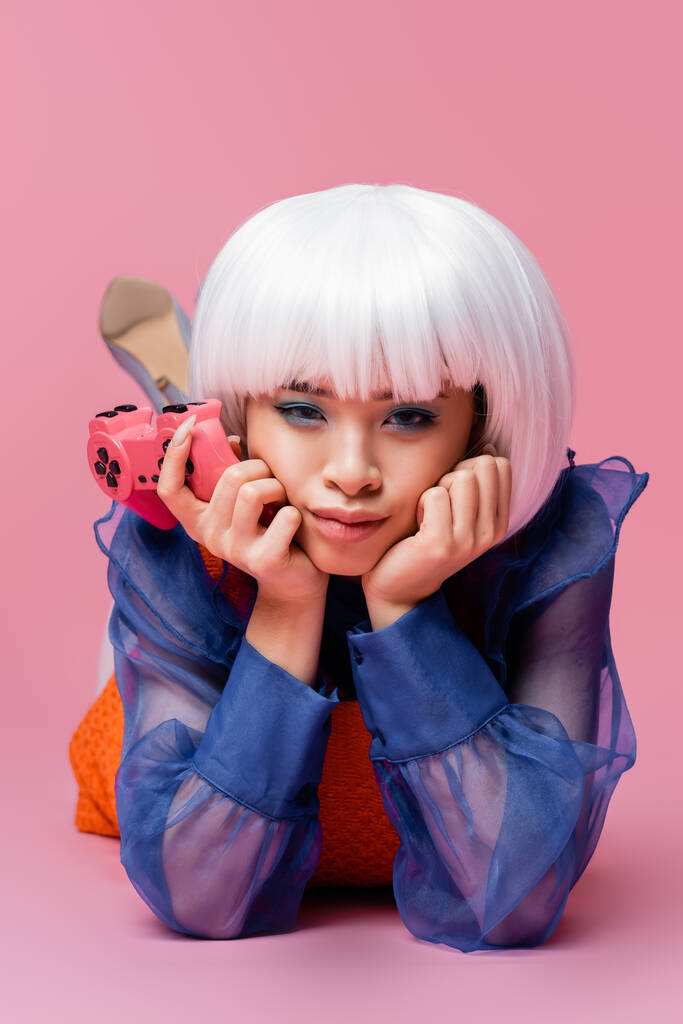 KIEW, UKRAINE - 10. DEZEMBER 2020: Asiatisches Pop-Art-Modell in weißer Perücke mit Joystick auf rosa Hintergrund liegend - Foto, Bild