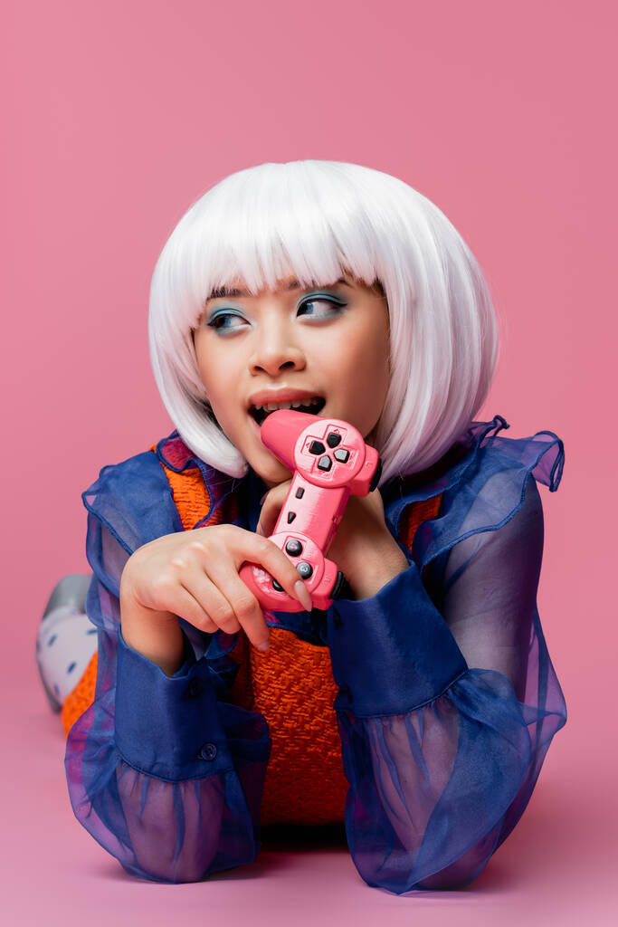 KYIV, UKRAINE - DECEMBER 10, 2020: Young asian woman in white wig biting joystick on pink background - Photo, Image