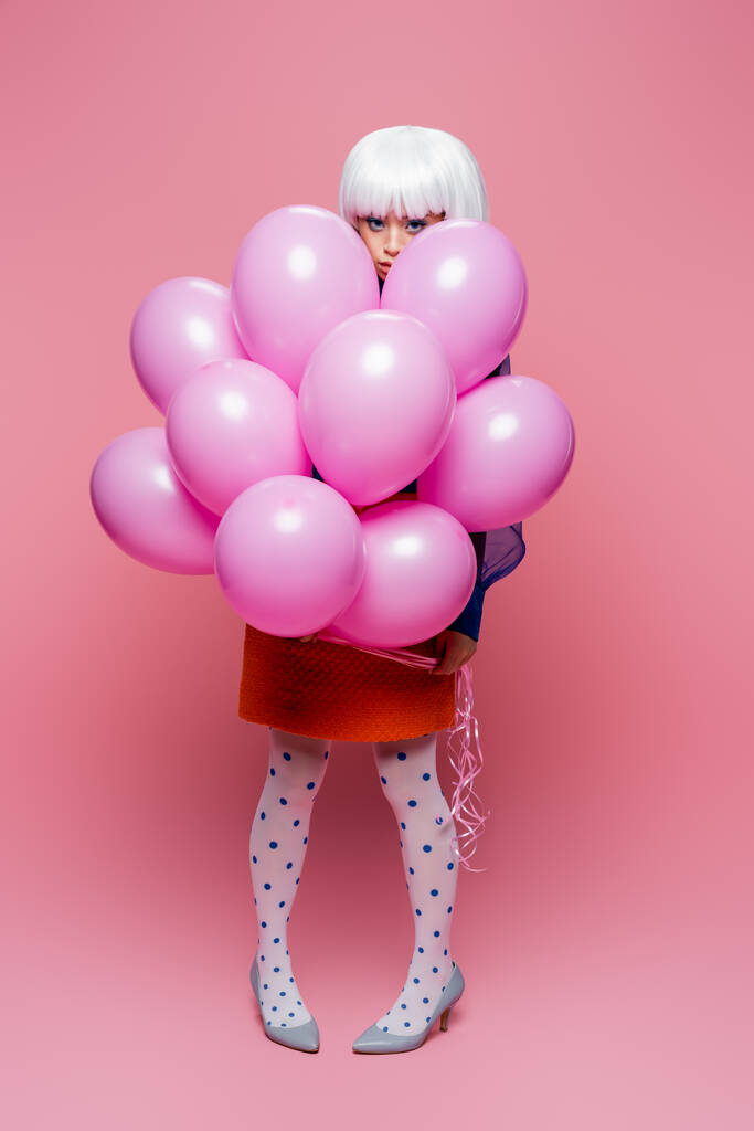 Aziatisch pop art model in hakken op zoek naar camera in de buurt ballonnen op roze achtergrond - Foto, afbeelding