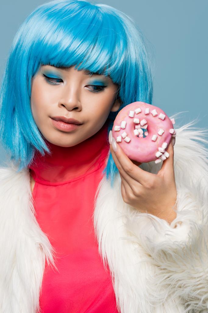 Portret van stijlvolle aziatische vrouw op zoek naar donut met glazuur geïsoleerd op blauw  - Foto, afbeelding