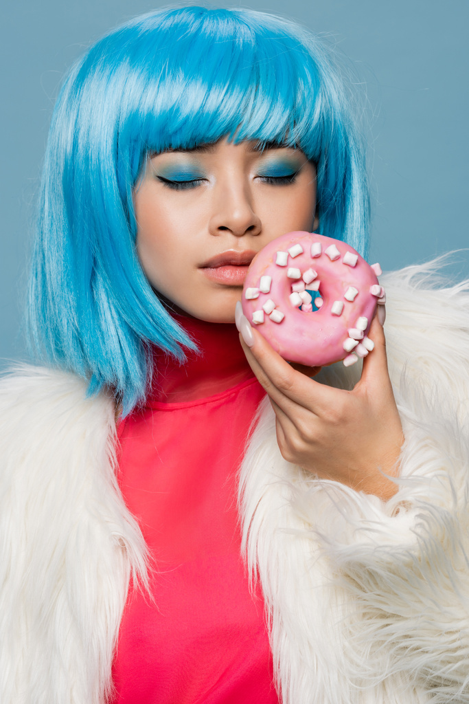 aziatische vrouw in pop art stijl sluiten ogen terwijl het houden van smakelijke donut geïsoleerd op blauw  - Foto, afbeelding