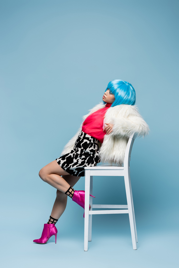 Kabarık ceketli, parlak peruklu, mavi arka planda sandalyenin yanında poz veren şık bir kadın.  - Fotoğraf, Görsel
