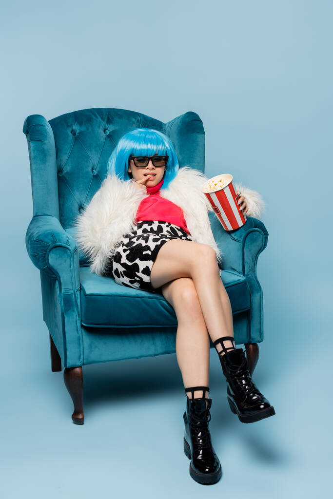 Stylish asian woman in fluffy jacket and sunglasses holding popcorn in armchair on blue background  - Photo, Image
