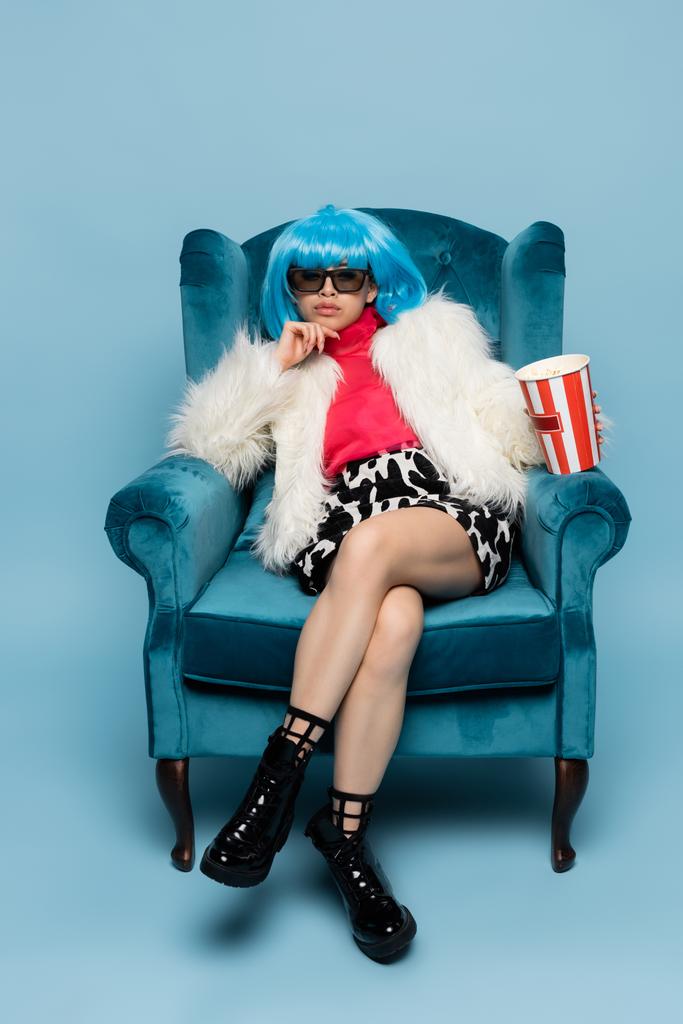 Asijské žena v pop arty stylu držení popcorn zatímco sedí na velveteen křeslo na modrém pozadí  - Fotografie, Obrázek
