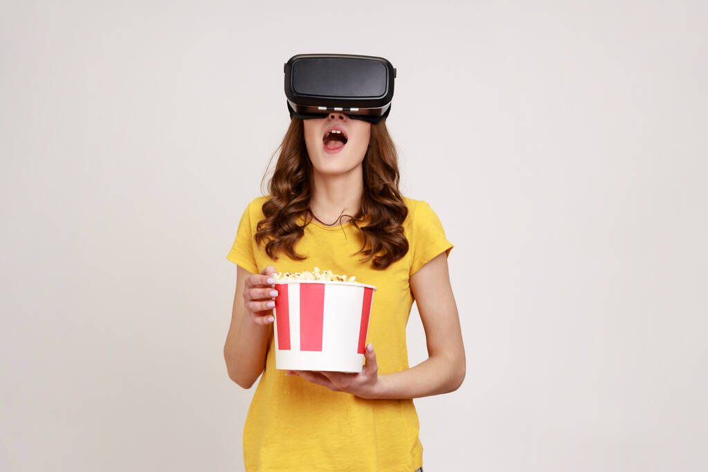 Giovane ragazza in giallo T-shirt ottenere esperienza VR auricolare, utilizzando occhiali realtà aumentata essere in realtà virtuale, guardando film con popcorn. Indoor studio girato isolato su sfondo grigio. - Foto, immagini