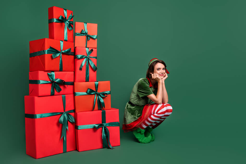 Foto de dulce soñadora joven elfo traje de cabeza sentado cerca de regalos montones de brazos mejillas sonriendo aislado color verde fondo - Foto, Imagen