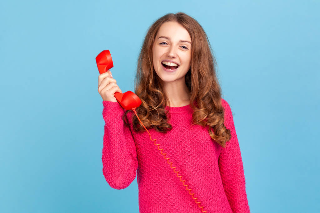 Портрет позитивной женщины в розовом пуловере, говорящей по стационарному телефону, держащей в руке телефон, смотрящей в камеру с зубастой улыбкой. Крытая студия снята на голубом фоне. - Фото, изображение