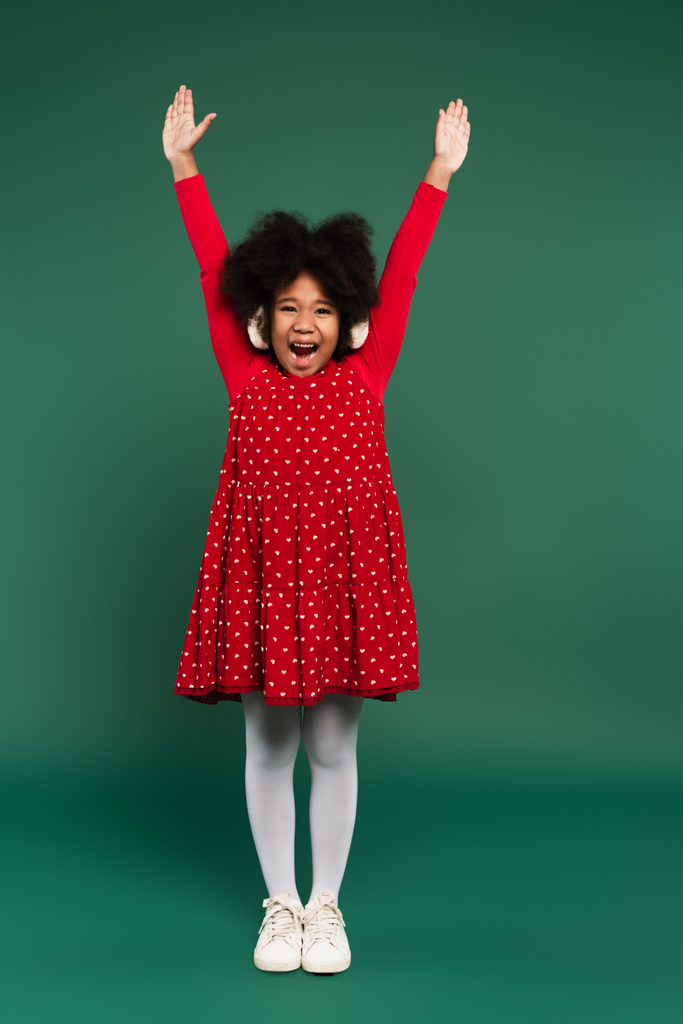 Vrolijk Afrikaans amerikaans kind in oor moffen en rode jurk het verhogen van de handen op groene achtergrond  - Foto, afbeelding