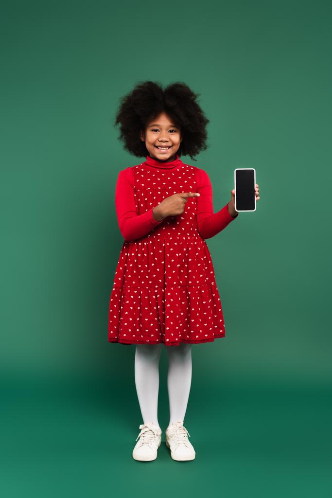 Sorridente ragazza afroamericana in abito che punta a smartphone con schermo bianco su sfondo verde  - Foto, immagini